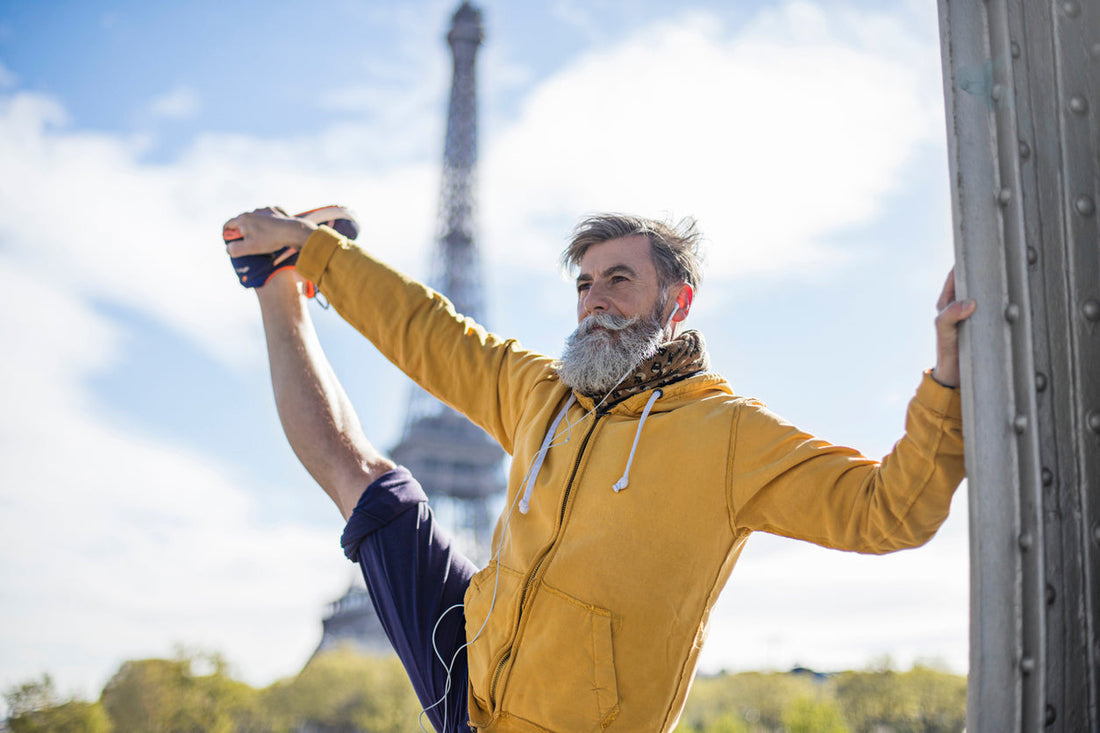 Alter bärtiger Mann in gelber Jacke streckt sein Bein hoc und hält es mit der rechten Hand fest.