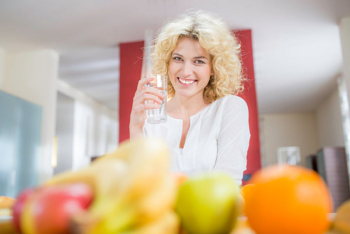 Eine blonde Frau die vor einem Tisch voller Obst ein Glas Wasser trinkt.