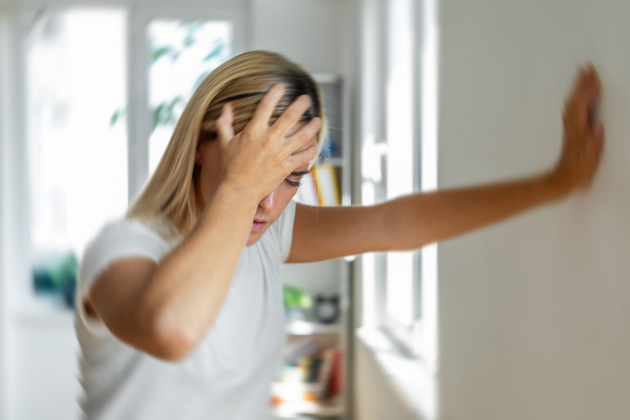 Blonde Frau hält sich aufgrund starker Migräne mit der rechten Hand den Kopf und mit der linken hand und stützt sich mit der linken hand an der Wand ab.