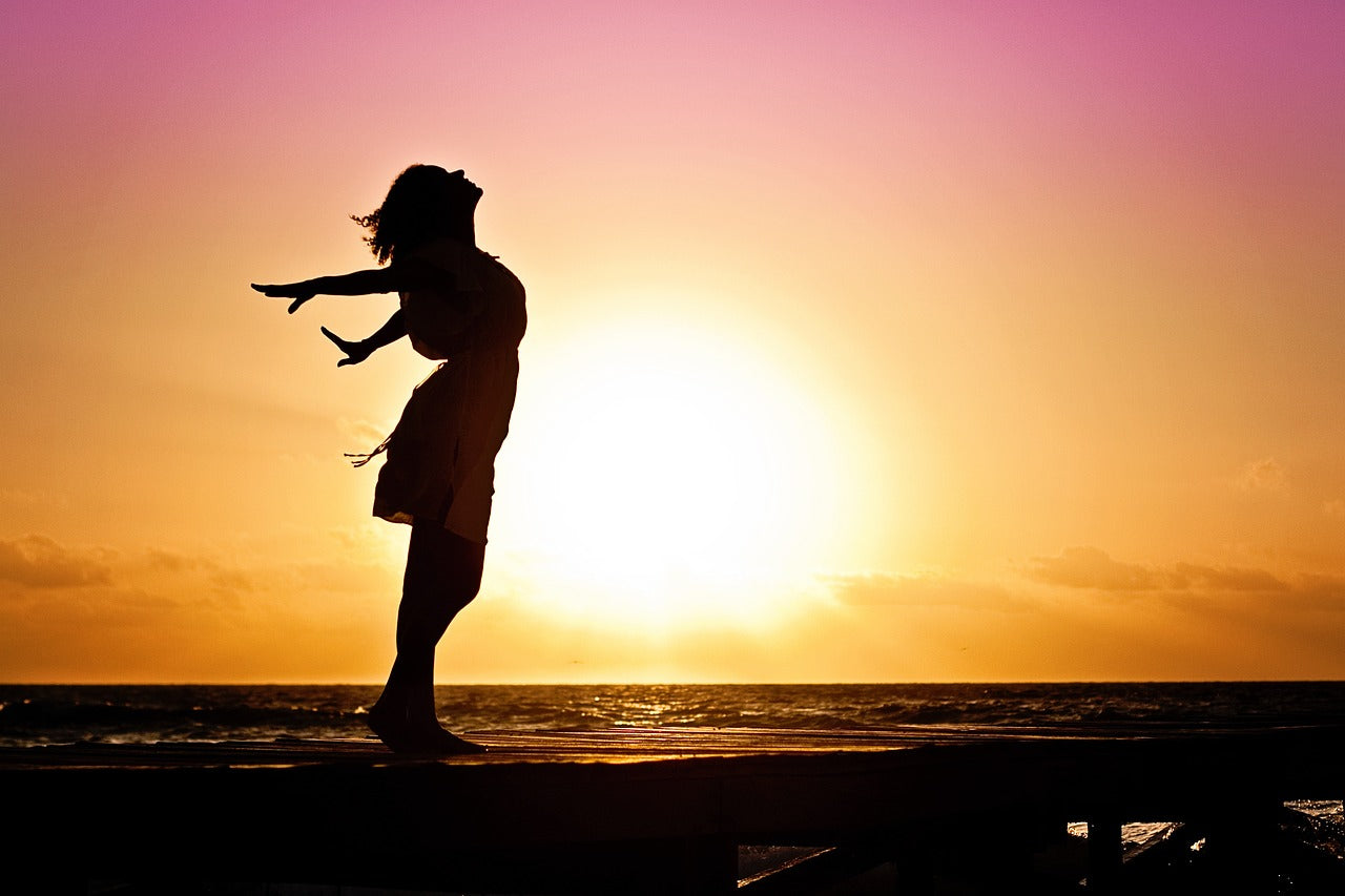 Eine Frau steht bei Sonnenuntergang am Meer und streckt den Kopf und ihre Arme nach hinten um tief einzuatmen.